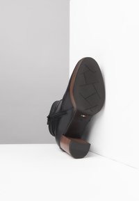 Stivale alla caviglia in pelle nera con zip laterale, tacco in legno marrone e suola in gomma texturizzata. Costruzione leggera, forma slanciata.