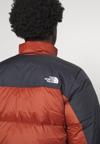 Pufová bunda v černé a oranžové barvě, s kostkovaným vzorem na spodní polovině, vysokým límcem a logem The North Face na rameni.