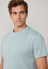 Tričko s krátkým rukávem, světle modré barvy, s texturovaným pruhovaným vzorem a kulatým výstřihem. Tkanina vypadá měkce a lehce.