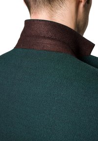 Zielony, teksturowany materiał z ciemnobrązowym kołnierzem. Zbliżenie na tył odzieży, podkreślające przeszycia kołnierza oraz kontrast kolorystyczny.