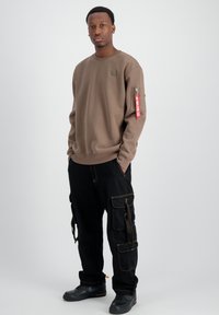Felpa marrone con logo frontale e tasca laterale con zip, abbinata a pantaloni cargo neri con multiple tasche e cuciture gialle.