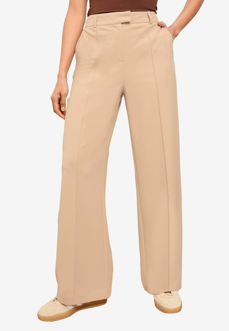 Pantalon large beige en tissu doux, avec taille haute, plis devant et poches sur les côtés, associé à des baskets de couleur claire.