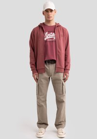 Bordo zip-up džemperis su bordo grafine marškinėliais, derinamas su šviesiais khaki krovinių džinsais ir kreminiais batais, papildytas baltu kepurėle.