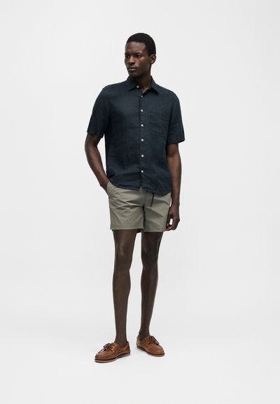 Άντρας που στέκεται και φοράει σκούρο πουκάμισο με κοντά μανίκια και κουμπιά, ανοιχτό γκρι σορτς και καφέ δερμάτινα παπούτσια σκαφών μπροστά από απλό φόντο.