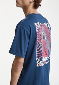 Ung mand i blå T-shirt med farverigt grafisk tryk af en siddende person og bølgemønstre på ryggen, der står sidelæns mod en hvid baggrund.