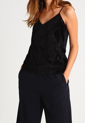 Kobieta w czarnej bezrękawnikowej bluzce z dekoltem w serek i luźnych czarnych spodniach, stoi z rękami w kieszeniach.