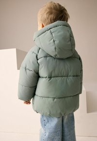 Steppjacke in sanftem Grün, mit großen gesteppte Bereichen und Kapuze. Kombiniert mit hellblauen Jeans, ist das Design schlicht und funktional.