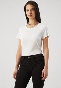 T-shirt bianca a maniche corte in tessuto leggero, abbinata a jeans neri aderenti con accenti in metallo dorato e una texture liscia.