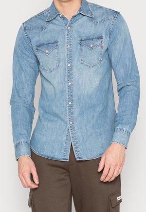 Mann trägt hellblaues Jeanshemd mit zwei Brusttaschen und braune Hose mit Cargotaschen.