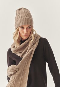 Knitattu beige-pipo, jossa on teksturoitu kuvio, yhteensopiva huivi ja käsineet. Malli pukeutuneena tumman väriseen yläosaan, esitellen pehmeitä materiaaleja ja lämpimiä sävyjä.