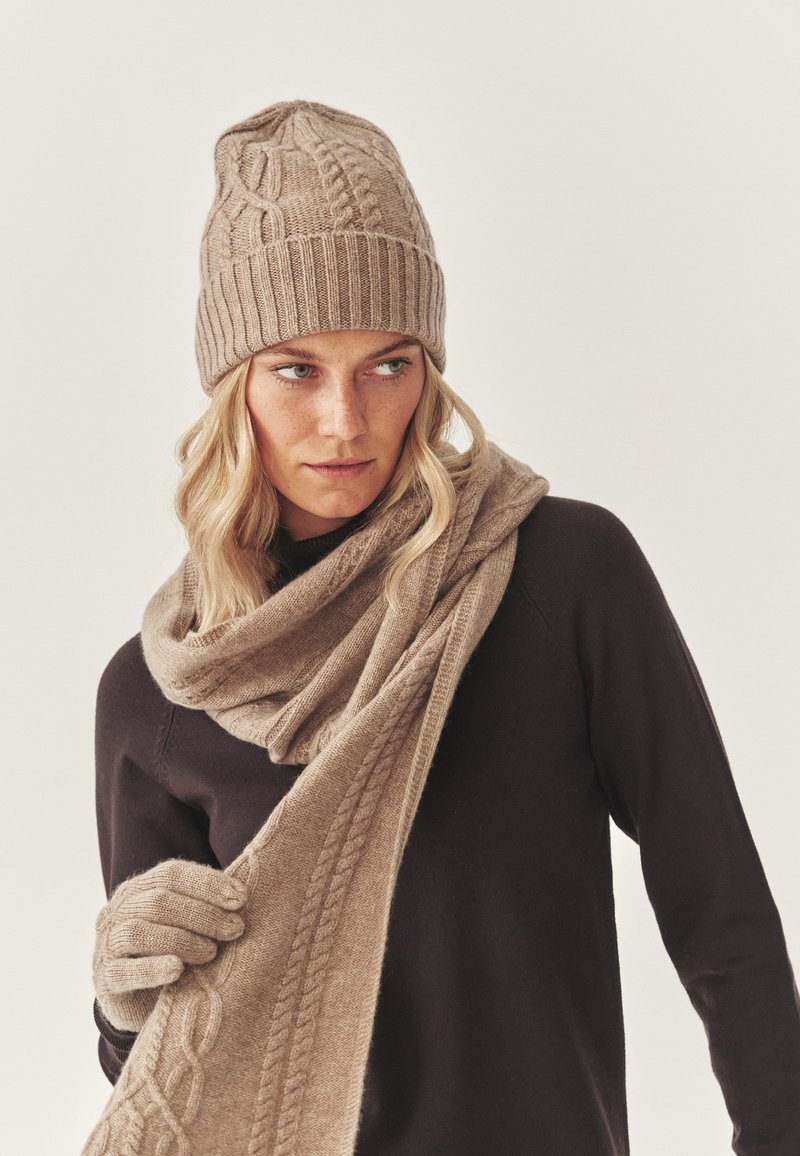 Knitattu beige-pipo, jossa on teksturoitu kuvio, yhteensopiva huivi ja käsineet. Malli pukeutuneena tumman väriseen yläosaan, esitellen pehmeitä materiaaleja ja lämpimiä sävyjä.