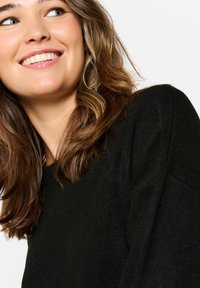 Un maglione nero presenta in modo prominente un design a maglia testurizzata. Il modello ha i capelli castano ondulati e un sorriso ravvicinato, con lineamenti delicati.