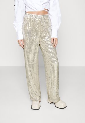 Witte cropped blouse met lange mouwen gecombineerd met hooggetailleerde, brede goudkleurige paillettenbroeken en beige plateauzolen.