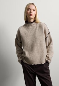 Pull-over beige en maille douce avec des accents marron foncé le long des poignets et de l'ourlet. Associé à un pantalon marron foncé.