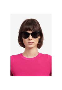 Occhiali da sole neri a forma di occhi di gatto con lenti scure, caratterizzati da una finitura lucida. Indossati da una persona con una maglia aderente a costine di un vivace colore rosa.