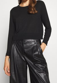 Donna che indossa un maglione nero a maniche lunghe e pantaloni larghi in pelle nera a vita alta, con una mano in tasca.
