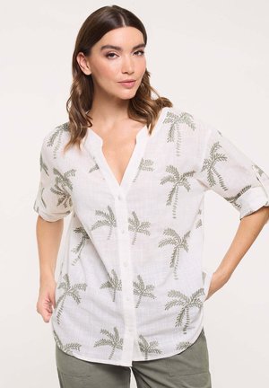Mujer con el cabello castaño hasta los hombros, con una camisa blanca abotonada con estampados de palmeras verdes y pantalones verdes, de pie con las manos en las caderas.