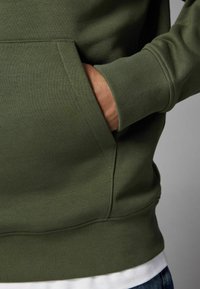Haut à capuche vert olive avec des poignets et un ourlet côtelés. Présente une poche avant et une texture douce en mélange de coton.