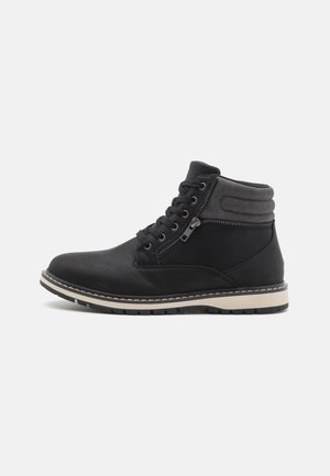Botas de tobillo negras hechas de material sintético texturizado, con un cierre de cordones en la parte delantera y una cremallera lateral, y una suela de goma blanca en contraste.