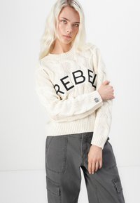 Suéter de malha em creme com "REBEL" em letras pretas em negrito no peito, combinado com calças cargo cinzas com bolsos laterais.