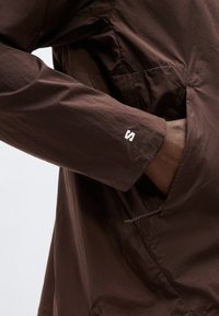 Μανίκι σκούρου καφέ μπουφάν με λευκό λογότυπο "S" στον καρπό· το χέρι μερικώς μέσα σε πλαϊνή τσέπη με ορατό φερμουάρ.