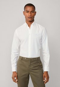 Camisa blanca de manga larga hecha de algodón, con cuello y un logo azul claro. Combinada con pantalones caqui.