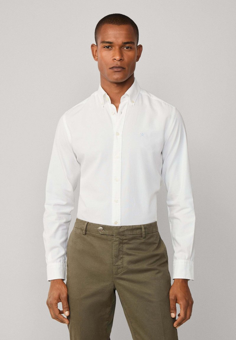Camisa blanca de manga larga hecha de algodón, con cuello y un logo azul claro. Combinada con pantalones caqui.