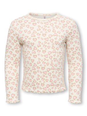 Jeune fille aux cheveux bouclés en pyjama rose clair à motifs de cœurs, souriante et levant une main sur un fond uni.