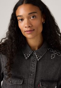 Chemise en denim noir avec un col pointu, des embellissements à clous sur les épaules et des fermetures à boutons avec deux poches avant.