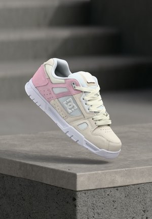 Weißer und rosa DC-Sneaker mit Schnürsenkeln schwebt über einem Betonsims mit Treppen im Hintergrund.