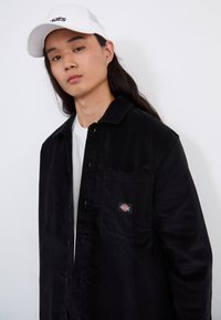 Camisa de pana negra con un bolsillo en el pecho y una etiqueta de marca, usada sobre una camiseta blanca. Cuenta con un cuello con solapa y cierre de botones.