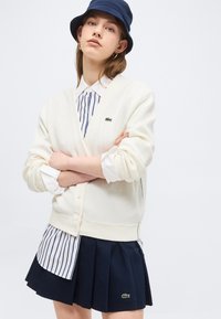 Jeune femme portant un chapeau seau bleu marine, un cardigan blanc boutonné sur une chemise rayée, et une jupe plissée bleu marine avec des logos de crocodile, debout les bras croisés.