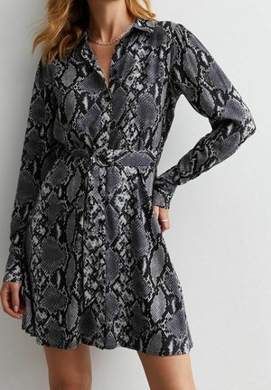 Žena v krátkých šatech s dlouhými rukávy, páskem, vzorem hadí kůže v černé a šedé barvě a s límečkem.