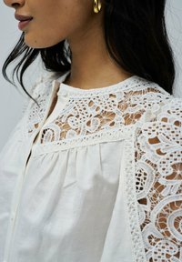Blusa blanca con intrincados detalles de encaje en los hombros y el escote, con cierre de botón y tejido suave y ligero.
