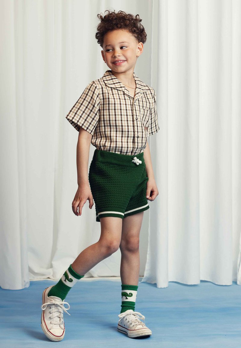 Short-sleeve, plaid button-up shirt in beige and brown paired with green knit shorts featuring white trim and a decorative drawstring. Worn with ankle socks and sneakers.