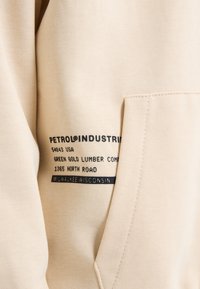 Sudadera de algodón beige con un bolsillo frontal y detalles de marca impresos en negro en el lateral, que incluyen la ubicación y la información de la empresa.