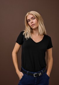 Blondýnka s vlasy po ramena, oblečená v černém tričku s výstřihem do V a modrých džínách s černým páskem, stojící s rukama v kapsách.