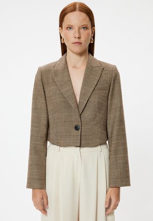 Korte bruine geruite blazer met een notch-kraag, sluiting met één zwarte knoop en lange mouwen, gecombineerd met crèmekleurige broeken.