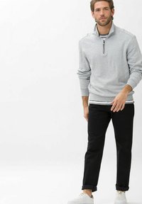Graue Zip-Hoodie mit geripptem Saum, über einem gestreiften Shirt getragen. Kombiniert mit schwarzen, hochgekrempelten Hosen und weißen Sneakers.