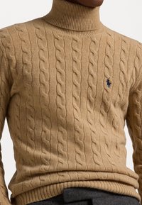 Beżowy sweter z golfem w splot kratkowany, z prążkowanymi mankietami i dolnym brzegiem; na piersi ma małe niebieskie logo. Miękka tekstura i dopasowany krój.