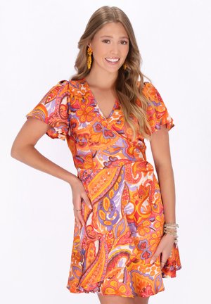 Ung kvinde, der smiler, iført en farverig orange, lilla og lyserød blomster-paisley kjole med øreringe og armbånd, foran en hvid baggrund.