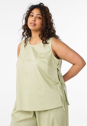 Žena s kučeravými vlasmi, ktorá nosí svetlozelený bezrukávový top so zaväzovaním na bokoch a voľné nohavice na mieru, stojaca s rukou za chrbtom.