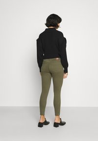 Suéter preto, com textura em costura canelada e ombros caídos, combinado com calças justas em verde-oliva. O conjunto apresenta um design de cintura baixa e mules com salto bloco.