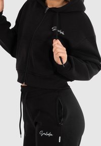 Felpa nera corta con zip, dotata di una tasca frontale e logo bianco. Abbinata a pantaloni jogger coordinati con vita elasticizzata.