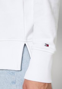 Sudadera blanca con puños acanalados y una abertura lateral. Incluye un pequeño logo en rojo, negro y blanco cerca del puño. Textura suave.