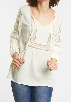 Vrouw draagt een witte blouse met kanten panelen en trekkoorden, gecombineerd met blauwe jeans, getoond van schouders tot heupen.