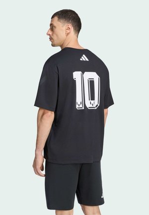 Mann trägt schwarzes Sportshirt mit weißer Nummer 10 und Adidas-Logo, kombiniert mit schwarzen Shorts, seitlich stehend vor hellem Hintergrund.