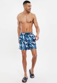 Pantaloncini da nuoto blu e bianchi con motivo a foglie, dotati di vita elastica e cordoncino. Indossate un paio di sandali blu.
