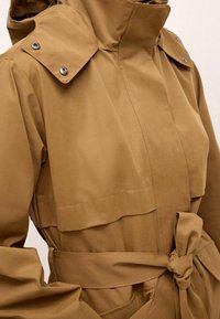 Khaki trenchcoat met een hoge kraag, knopenepauletten en een ceintuur op de taille. Beschikt over een gladde stof en voorzakken.