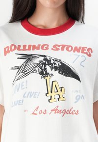 White t-shirt with red collar featuring a graphic of an eagle, "Rolling Stones," "LA," and concert details in various colors.